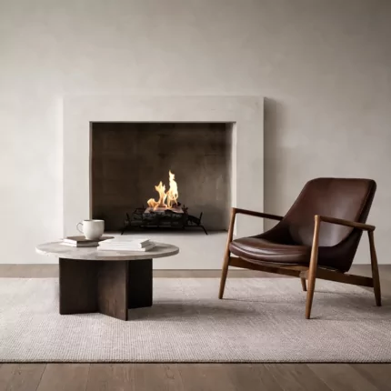 Modern minimalist living room featuring a built-in fireplace with a clean-burning flame set in a recessed concrete surround. In front, a round stone coffee table with books and a white mug sits on a textured rug beside a mid-century style brown leather lounge chair with wooden arms, creating a warm, contemporary interior setting.