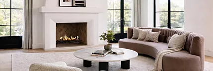 EcoSmart Fire Heritage 50 Grate installed in a white built-in fireplace with visible flame, set in a bright contemporary living room featuring a curved sofa, marble coffee table and large black-framed windows.
